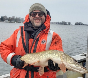 A light-skinned man with a beard wears coldwater safety gear, gloves, a knit cap, and sunglasses, standing on a boat in a river, holding up a large speckled fish