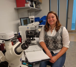 A light-skinned woman wearing a sweater vest smiles and sits next to a microscope in a lab with a binder open on the counter next to her