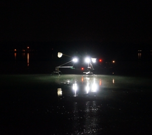 a boat at nighttime with bright headlights and two booms extended over the water, and two people with nets at the front