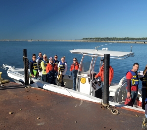 a group of people stand on a fiberglass boat with a bimini top at dock. Two people are on the dock holding or tying the lines to the dock