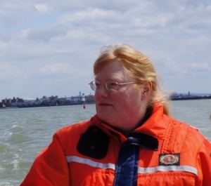 A ruddy light-skinned woman with reddish blond hair and glasses wears coldwater safety gear while standing behind a seat on a boat in a river