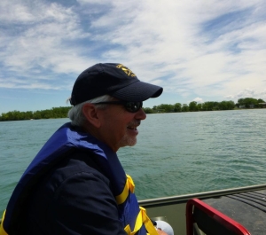 A light-skinned man in a hat and sunglasses wearing a life jacket sits on a boat in a river