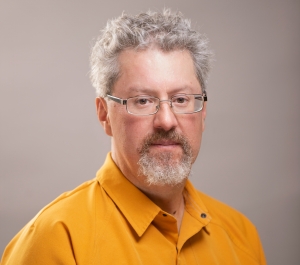 A ruddy light-skinned man with graying hair and beard wearing glasses and a yellow-orange buttoned shirt