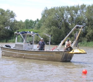 two people on a metal boat in a river use a boom to lift a large piece of equipment