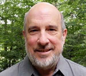 A smiling light-skinned balding man with gray hair and beard, wearing a grey buttoned shirt stands before some trees outside