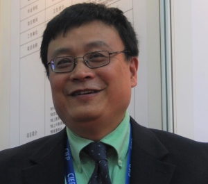 A light tan-skinned man with short dark hair and glasses wears a suit and tie and a lanyard