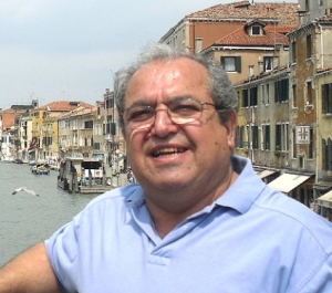 A light tan-skinned man with gray hair and glasses sits near some buildings along a canal