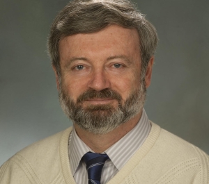 portrait of Alexander Karatayev, a light-skinned person with short graying hair and beard wearing a cream sweater over a striped collared shirt and blue striped tie