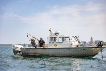 Two people on the back deck of a boat with a cabin on a lake by a city. One pulls on a rope over a pulley to pull in equipment from the water.