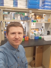 A person with short brown hair wearing a denim shirt takes a selfie in a lab