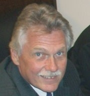 a person with graying hair and mustache and a gap-toothed smile in a suit and tie