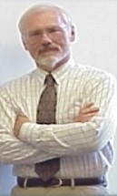 A light-skinned man with white hair and cropped beard and glasses standing with arms crossed while wearing a button-down shirt and tie