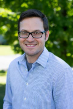 A person with glasses and short dark hair wearing a blue patterned button-down shirt smiles while standing outside