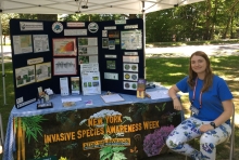 A person sits in a chair next to a table with an informational display outdoors. There is a large posterboard and a banner that says "New York Invasive Species Awareness Week."
