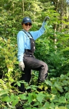 A person stands in the brush of a forest wearing a hat, sunglasses, overalls, and work gloves