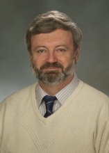 portrait of Alexander Karatayev, a light-skinned person with short graying hair and beard wearing a cream sweater over a striped collared shirt and blue striped tie