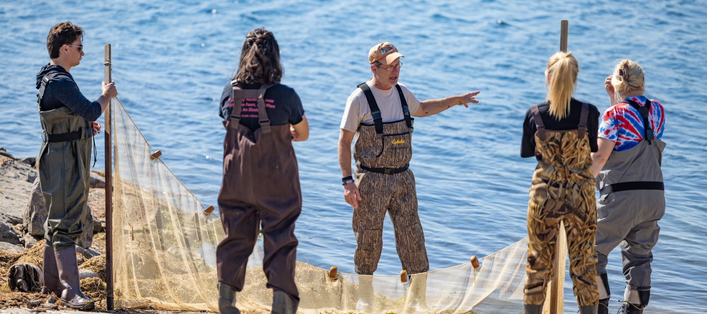 a class listens to an instructor explain how to use a seine net outdoors near the water, everyone wears hip waders