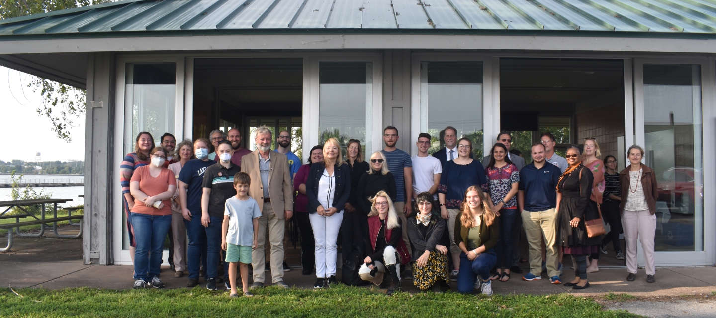 a group picture with about 30 people in front of a pavilion by the waterfront