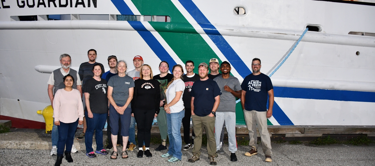 Fifteen people pose in two rose in front of a docked ship Lake Guardian