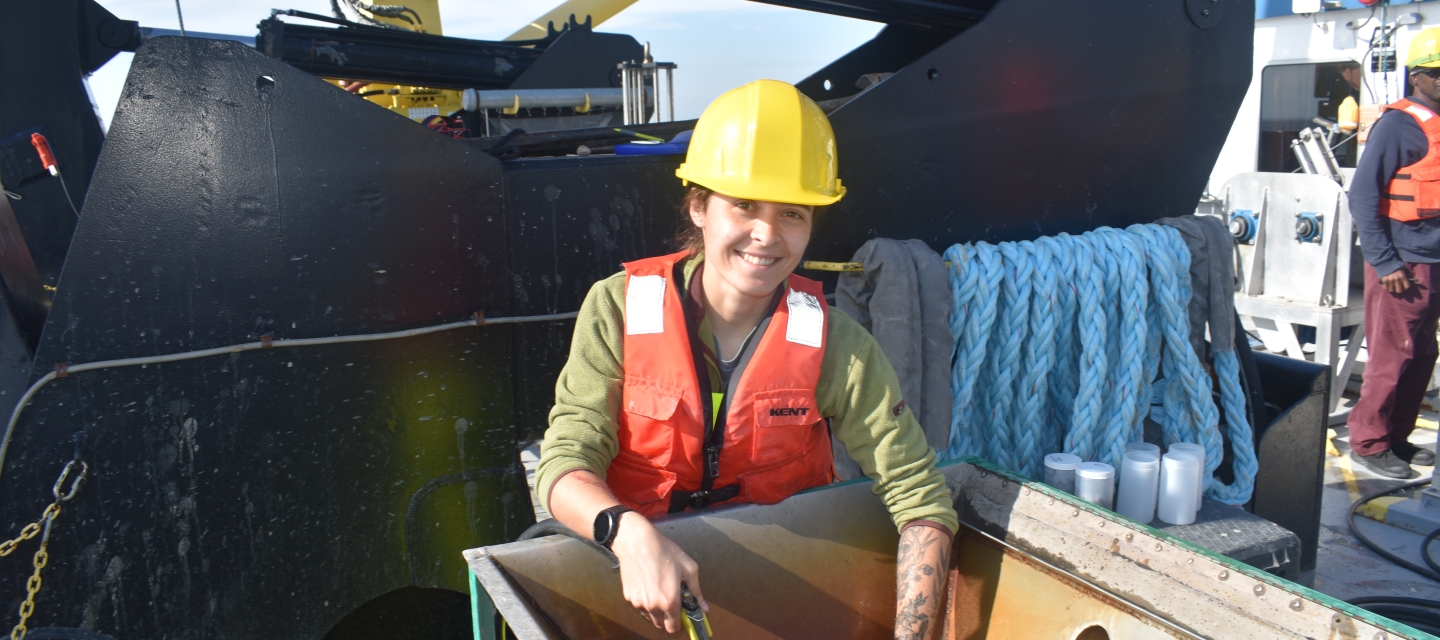 A light-skinned person in a life jacket and hard hat reaches into a basin of muddy water on the deck of a ship in front of a large hydraulic winch