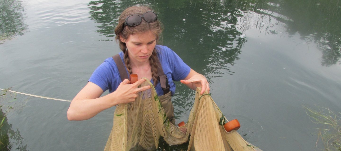 A light-skinned woman wearing chest waders stands in water up to her waist. She is holding and manipulating a net with floats on it.