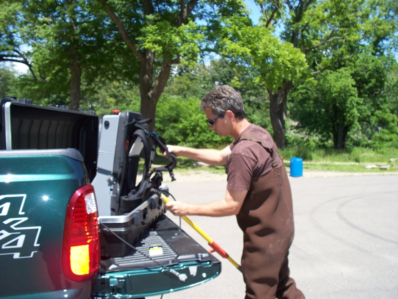 A person in chest waders takes a backpack unit and pole out of a case on the tailgate of a truck