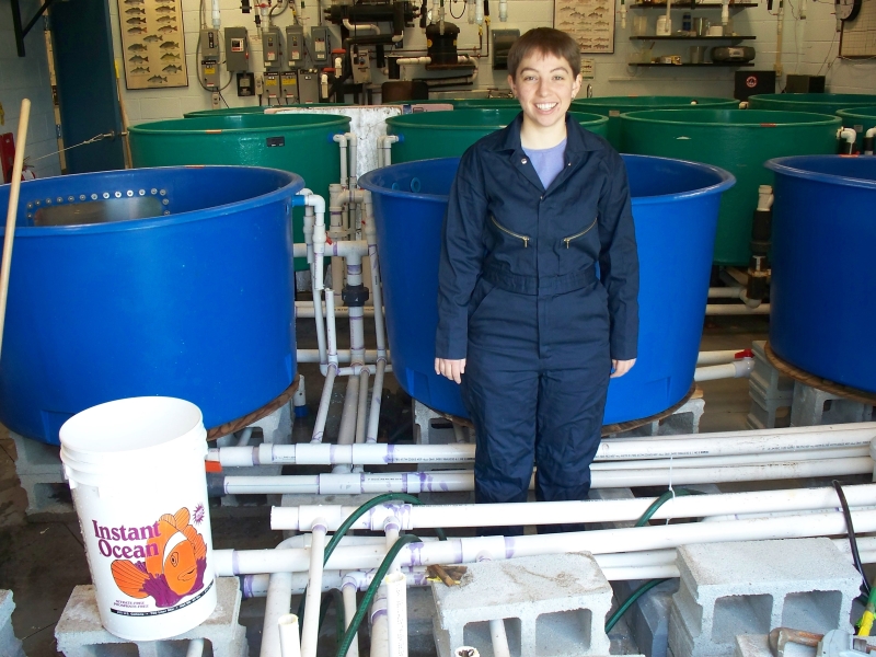 A person in coveralls poses in front of a row of large round tanks among exposed pipes and cinderblocks.