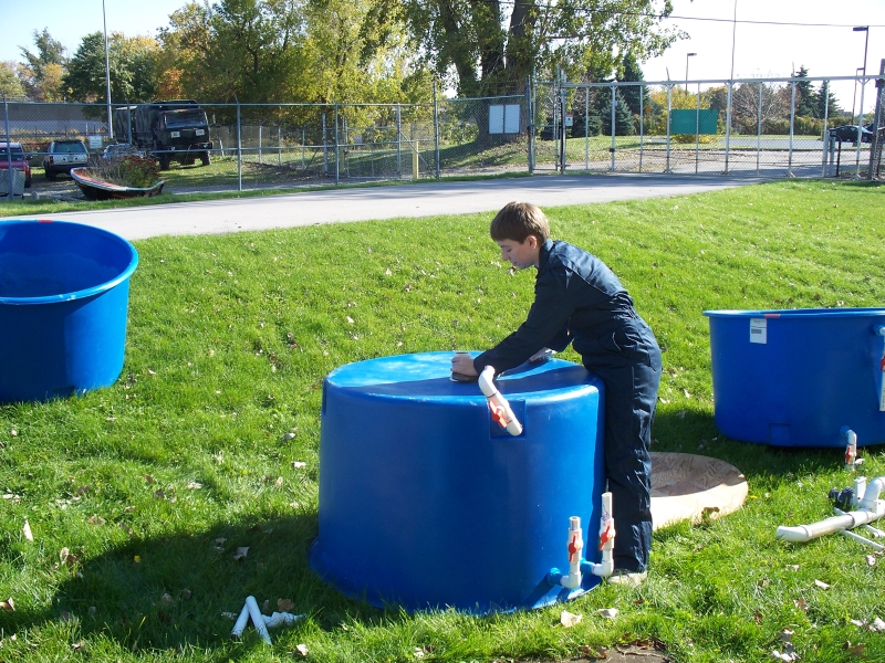 A person in coveralls removes a pipe from the bottom of a large round tank on the lawn. There are two other tanks and some pipe on the ground.