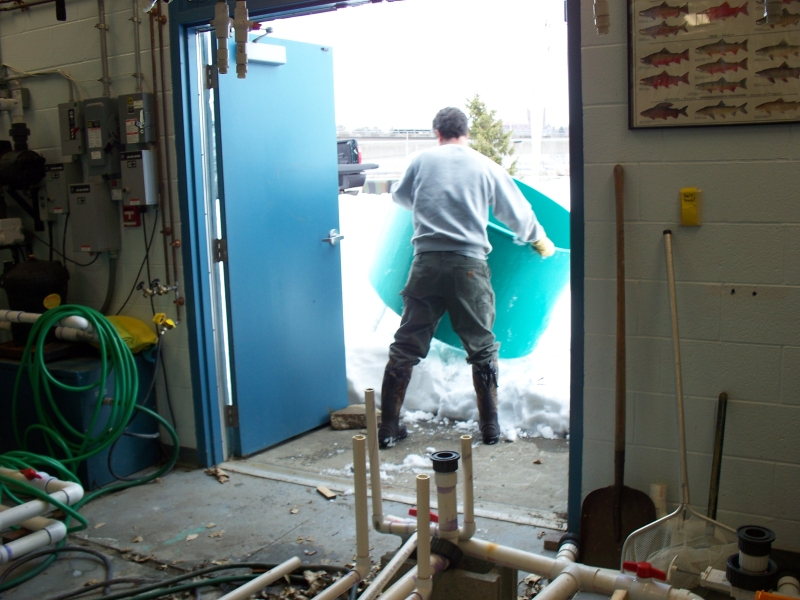 A person maneuvers a large round tank over a snow bank in the open doors of a room. There are exposed pipes on the floor.