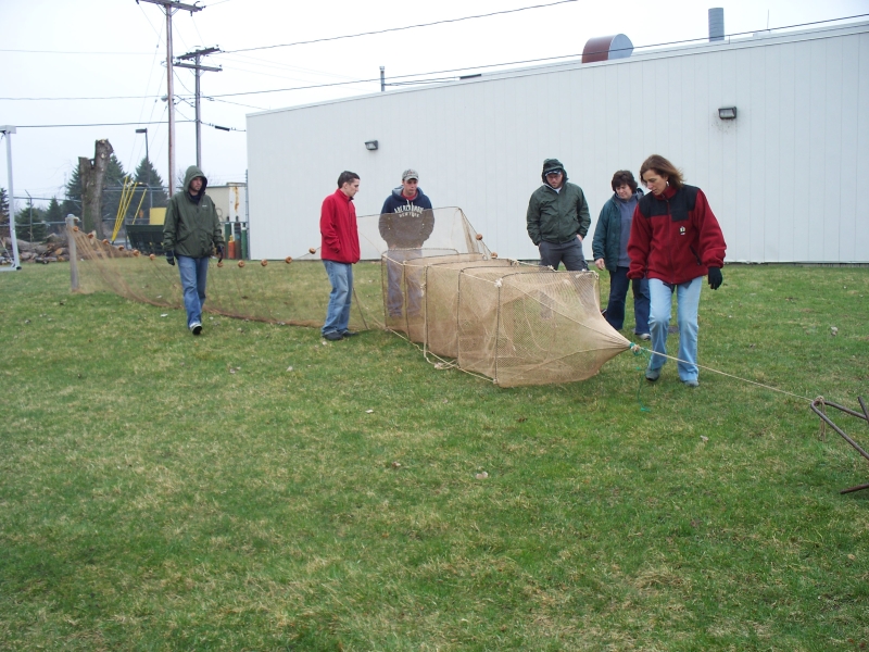 A group of people inspect a net set up on land. It has square frames, an anchor at one end, and a flat wing section coming off the front