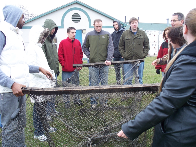 Ten people stand around and hold a fishing net open on a lawn. The net is box shaped and has two three-foot wooden beams as floats