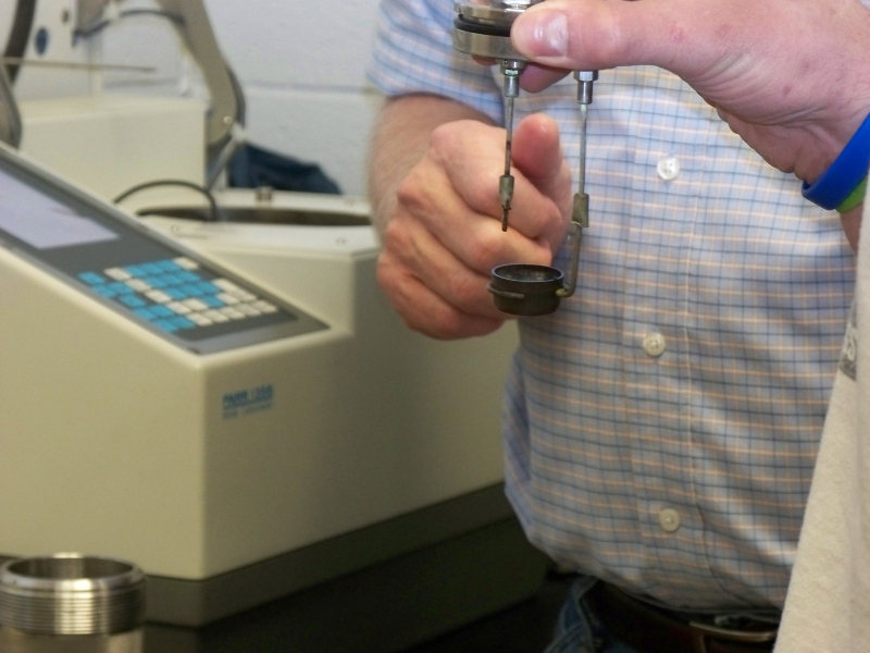 Closeup of someone's hands holding a metal lid with two rods hanging from it. At the bottom of one rod is a small metal cup. In the background is a lab instrument