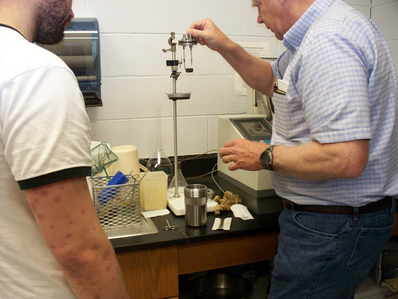 A person takes a metal lid with two rods and a tiny metal cup and prepares to set it on a stand. Nearby is a lab instrument and a large metal cup that the lid fits on. Someone watches.