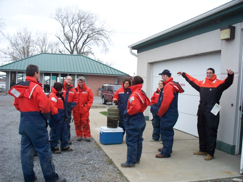 A group of people standing by a garage door wearing cold water safety gear. There is a cooler and some buckets.