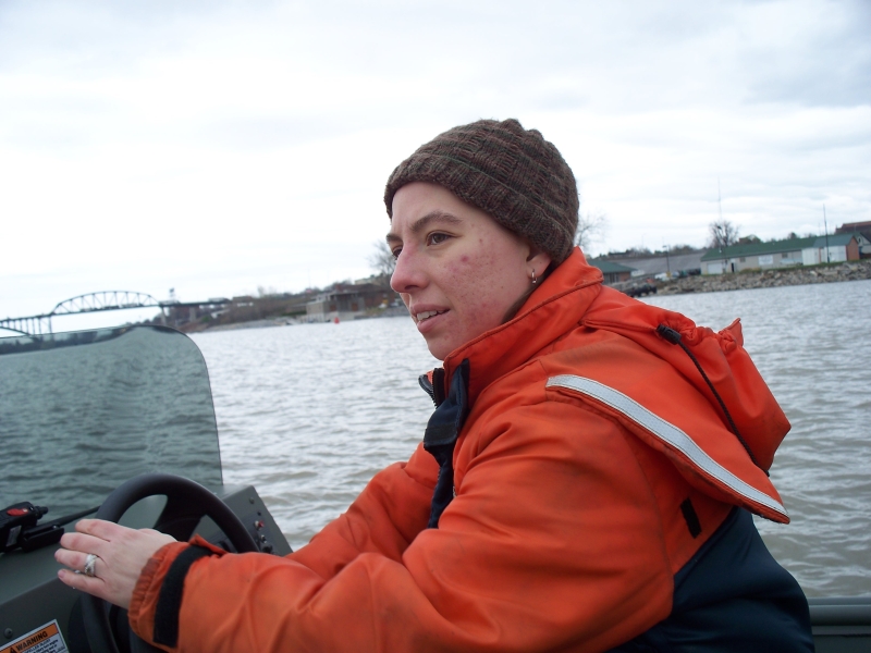 A person sitting at the helm of a boat, wearing coldwater safety gear and a knit cap