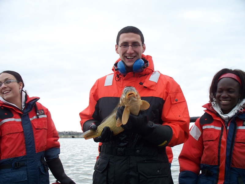 Three people wearing cold water safety gear by the water smile and laugh. The person in the middle holds up a carp