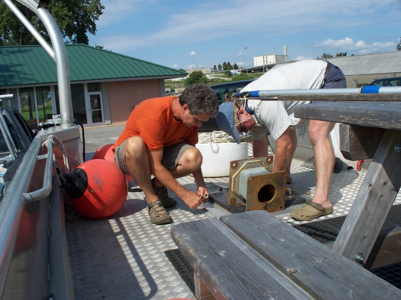 Two people kneeling to fasten something to the deck of a landing craft boat on a trailer in a parking lot