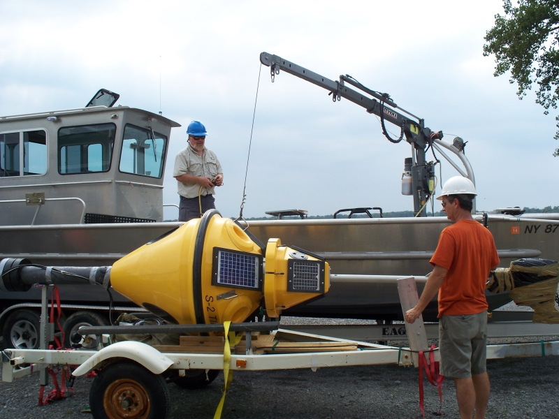 A person wearing a hard hat operates a small crane on the deck of a boat that's on a trailer in a parking lot. Another person supervises as the crane lifts a fiberglass buoy off a trailer.