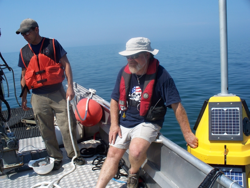 A person sits on the edge of a boat. A fiberglass buoy sits in the water next to them. Another person on the boat holds a rope.