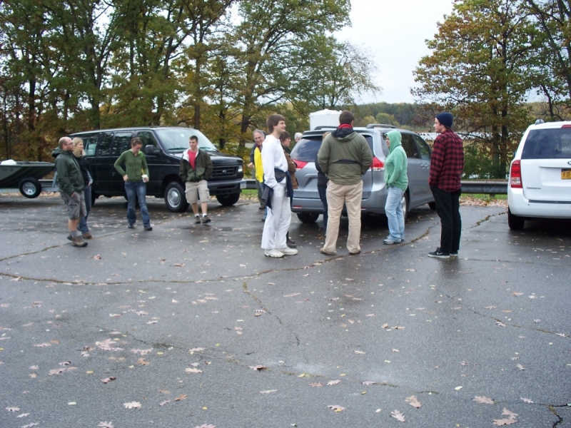 A group of people standing in a parking lot near some cars. A van is trailering a boat.