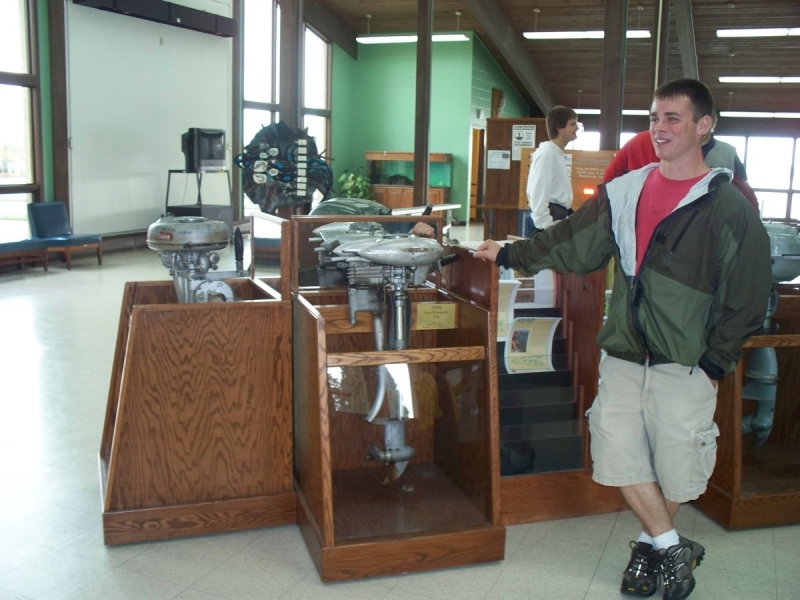 A few people stand near antique metal boat motors in wooden stands