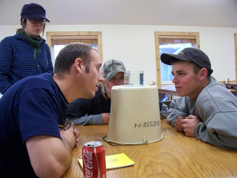 Four people look intently at a small cup of water set on top of an upside-down bucket. There are little white specks in the cup of water.