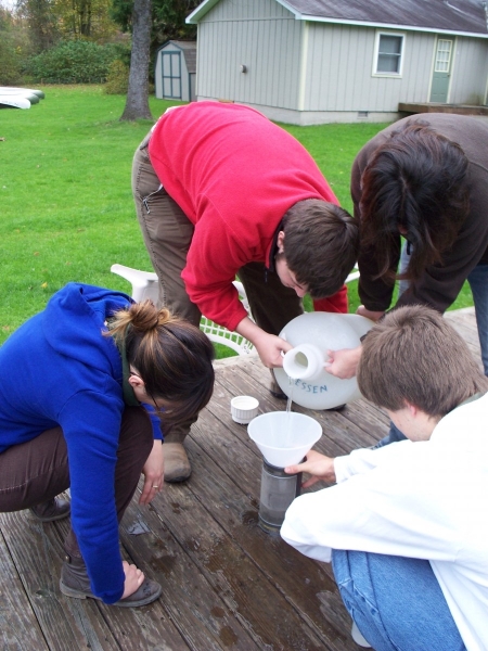 Four people work together to pour a jug of water into a funnel over a sieve on a deck outdoors.