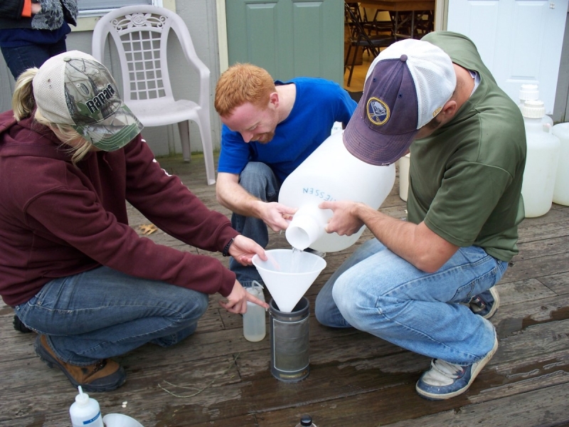 Three people work together to pour a jug of water into a funnel over a sieve, outdoors on a porch.