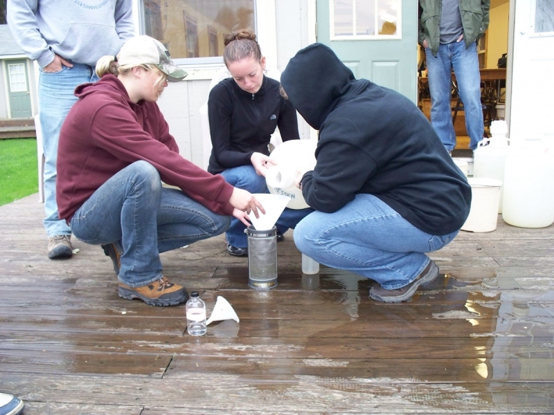 Three people work together to pour a jug of water into a funnel over a sieve, outdoors on a porch. Two people stand watching.