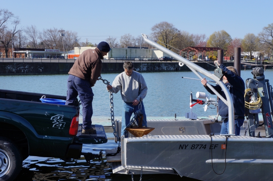 A person hands an anchor on a chain from the back of a pickup truck across to someone standing in the bow of a boat on a trailer. A third person adjusts a boom arm on the boat deck.