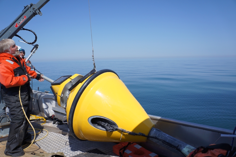 A buoy lays on the deck of a boat. A cable goes from a handle to a small crane. A person stands by the side near the controls of the crane.