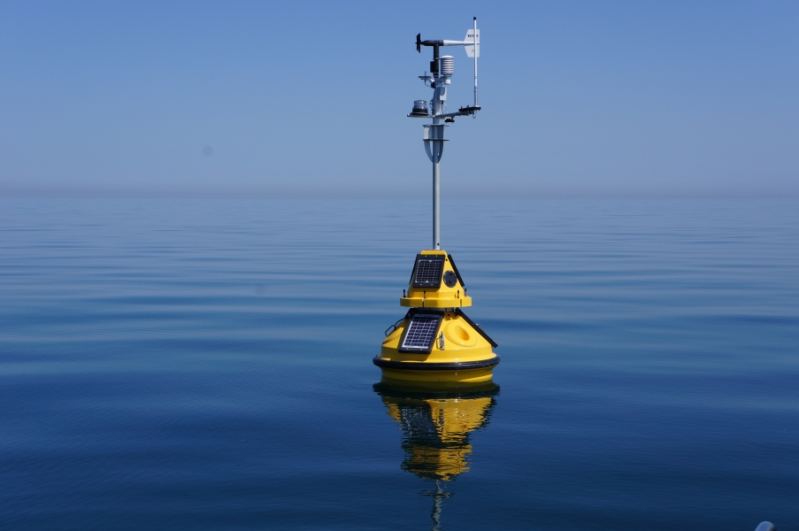 A science buoy with weather instruments on a tall mast floating on calm water. There is little deliniation between the clear sky and the calm water.