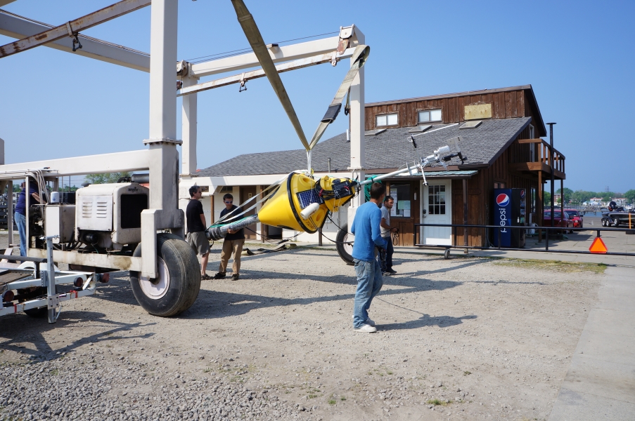 A buoy hangs from a strap in a large metal frame on wheels. Several people steady the buoy.