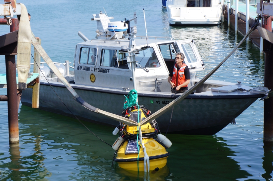 A person stands in the front of a boat called "R/V John J. Freidhoff," near a buoy in the water at the end of a boat slip. There are straps from the buoy to a metal frame.